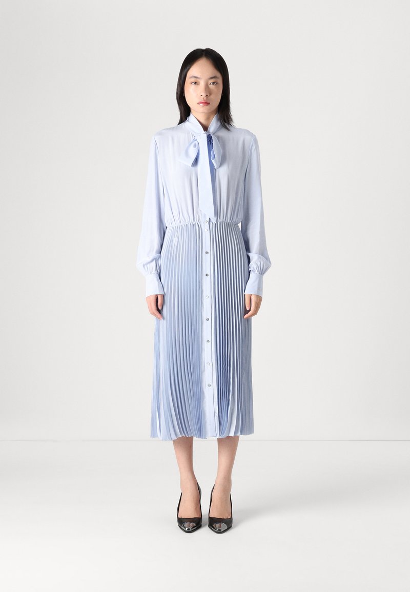 Vestido plissado azul claro com botões, mangas longas e detalhe de laço no pescoço. Combinado com sapatilhas pretas, com uma textura suave e um design fluido.