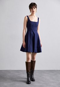 Robe bleu marine en A avec un décolleté carré et des bretelles larges. Une paire de bottes marron et noires jusqu'au genou avec une texture lisse et des matériaux mixtes.