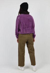 Un pull violet en peluche avec une finition texturée, associé à un pantalon vert olive. Le mannequin porte un bonnet noir et des baskets blanches.