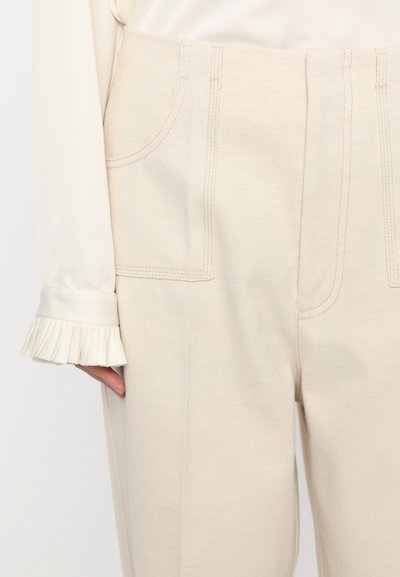 Pantaloni de culoare crem, cu o textură netedă, având două buzunare frontale și cusături proeminente. O manșetă din satin pliată este vizibilă.