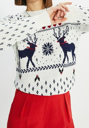 Vrouw met een witte kersttrui met donkerblauwe rendier- en sneeuwvlokpatronen, gecombineerd met rode, hooggetailleerde broek.