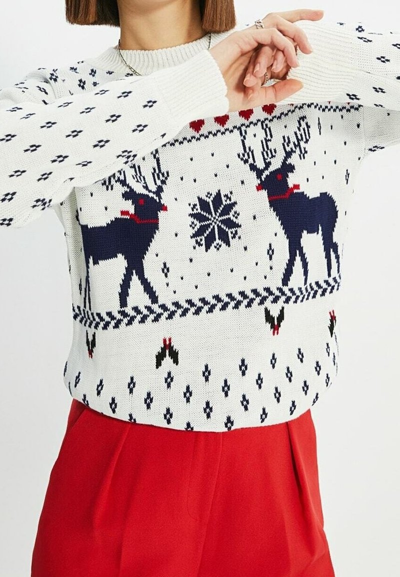 Femme portant un pull de Noël blanc avec des motifs bleu marine de rennes et de flocons de neige, associé à un pantalon rouge taille haute.
