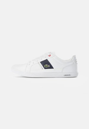 Baskets basses blanches avec une bande marine et un logo crocodile vert sur le côté, lacets blancs et marquage Lacoste sur la semelle et le talon.