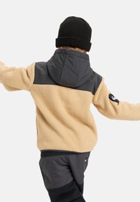 Chaqueta de forro polar beige con una sección superior y capucha en gris oscuro; logo negro en la manga. Combinada con pantalones oscuros, que muestran texturas suaves.