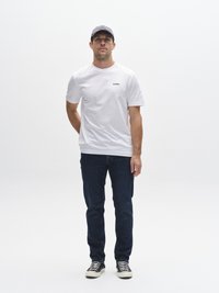 Camiseta blanca de manga corta con logo negro, combinada con jeans azul oscuro y zapatillas a rayas blancas y negras, complementada con una gorra gris.