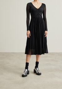 Černé skladované šaty s dlouhými průsvitnými rukávy a výstřihem do V. V páru se lesklými stříbrnými platformovými botami a černými žebrovanými ponožkami.