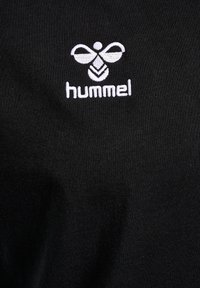 Zwarte stof met een gladde textuur, voorzien van een witte geborduurde logo van een gestileerde bij en het woord "hummel" eronder.