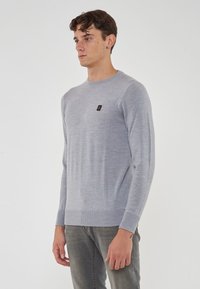 Maglione grigio chiaro realizzato in tessuto morbido, con un colletto a crew ribbed e polsini, dotato di un piccolo logo sul petto. Design semplice e aderente.