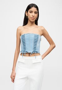 Γυναίκα με μακριά σκούρα μαλλιά φοράει αμάνικο γαλάζιο κορσέ με δαντελένιες λεπτομέρειες και λευκό ψηλόμεσο παντελόνι, μπροστά σε απλό φόντο.