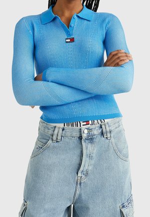 Kvinde med krydsede arme iført en blå ribbet langærmet polo og lyseblå denimjeans med synligt Tommy Jeans linning.