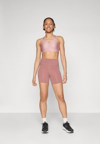 Sujetador deportivo de color rosa claro con cierre frontal, combinado con pantalones cortos de cintura alta a juego. Usado con zapatillas deportivas negras y calcetines blancos.