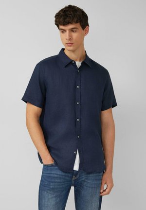 Man met kort donker haar, gekleed in een marineblauw overhemd met korte mouwen over een wit T-shirt en een blauwe spijkerbroek, staand tegen een effen grijze achtergrond.