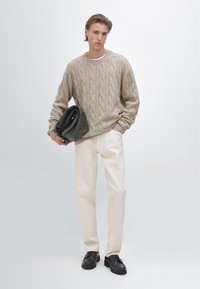 Pull beige en maille torsadée, pantalon crème et chaussures marron foncé. Le mannequin tient un sac gris, debout devant un fond clair.