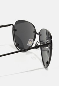 Lunettes de soleil noires avec une monture en métal, des verres ovales et des plaquettes de nez ajustables. Les verres sont gris foncé avec une surface lisse et réfléchissante.
