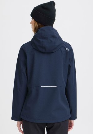 Kvinne iført navyblå hettejakke og svart lue, vist bakfra mot en ensfarget lys bakgrunn.