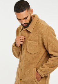 Chaqueta de forro polar color marrón claro con cuello, botones frontales y un bolsillo en el pecho. Presenta un acabado texturizado y bolsillos laterales para mayor funcionalidad.