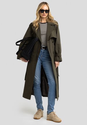 Olivgrüner Trenchcoat, gestreiftes schwarz-weißes Oberteil, blaue Jeans, beige Sneakers und schwarze Umhängetasche. Glatte Textur, langes Design, klassische Knöpfe.