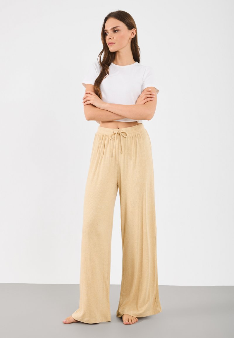 Beige wijde broek gemaakt van een zachte, gestructureerde stof met een elastische tailleband en een strik aan de voorkant. Gekleed met een kort mouwtjes wit top.