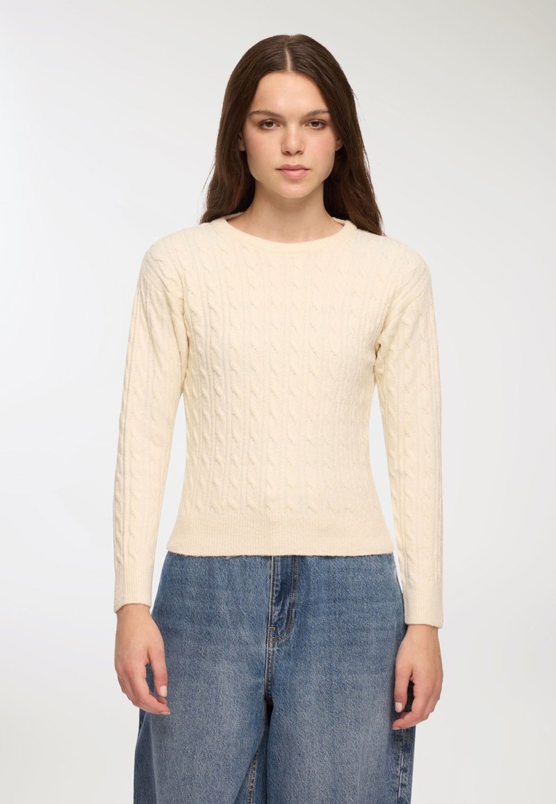 Młoda kobieta z długimi brązowymi włosami, ubrana w kremowy sweter w warkocz oraz luźne niebieskie dżinsy, stoi na jednolitym tle.