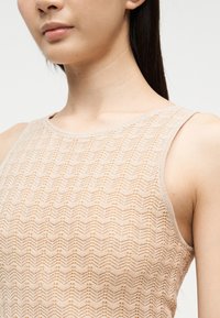 Κοντινό πλάνο ενός ατόμου που φοράει αμάνικο μπεζ πλεκτό τοπ με σχέδιο σε σχήμα κεφάλι βέλους, εμφανίζοντας τον λαιμό, τον ώμο και το πάνω μέρος του στήθους.