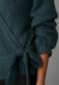 Teal gestrickter Pullover mit geripptem Muster, langen Ärmeln und einem Stoffgürtel, der an der Taille zu einer Schleife gebunden ist. Die Muster sind subtil und gemütlich.