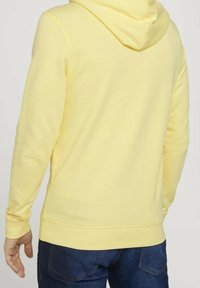 Felpa gialla di cotone con tasca a marsupio, polsini e orlo a costine. Dotata di cappuccio con coulisse e texture liscia. Vista da dietro.
