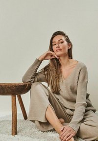 Beige ribbed knit sweater with V-neck, paired with matching wide-leg pants. Model seated beside a wooden table, on a plush white carpet.