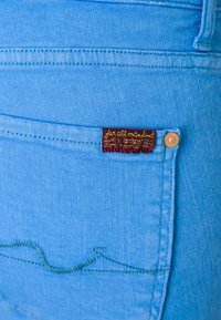 Jeans de mezclilla azul claro con una etiqueta en marrón que presenta texto dorado. Detalle de costura sobre el bolsillo trasero en un patrón ondulado.