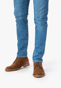 Braune Wildleder-Ankle-Boots mit Schnürung, ausgestattet mit einer hellbraunen Sohle und Akzenten. Getragen mit enganliegenden blauen Jeans.