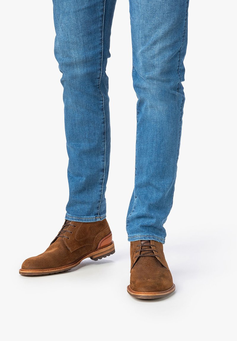 Braune Wildleder-Ankle-Boots mit Schnürung, ausgestattet mit einer hellbraunen Sohle und Akzenten. Getragen mit enganliegenden blauen Jeans.