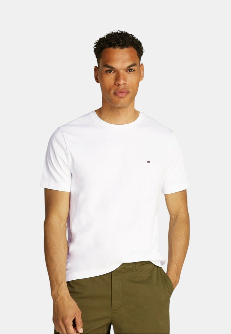 T-shirt bianco a maniche corte in cotone, con collo rotondo e un piccolo logo sul lato sinistro del petto. Abbinato a pantaloni verde oliva.