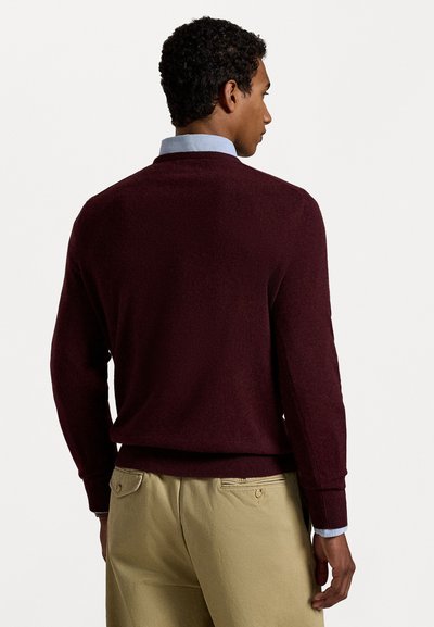 Suéter burdeos de corte ajustado, con mangas largas, puños y dobladillo de canalé. Llevado sobre una camisa de cuadro azul claro, con textura suave.