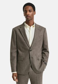 Giacca da abito a zigzag marrone con rever a punte, due tasche frontali e chiusura a un bottone. Indossata sopra una camicia di colore chiaro.