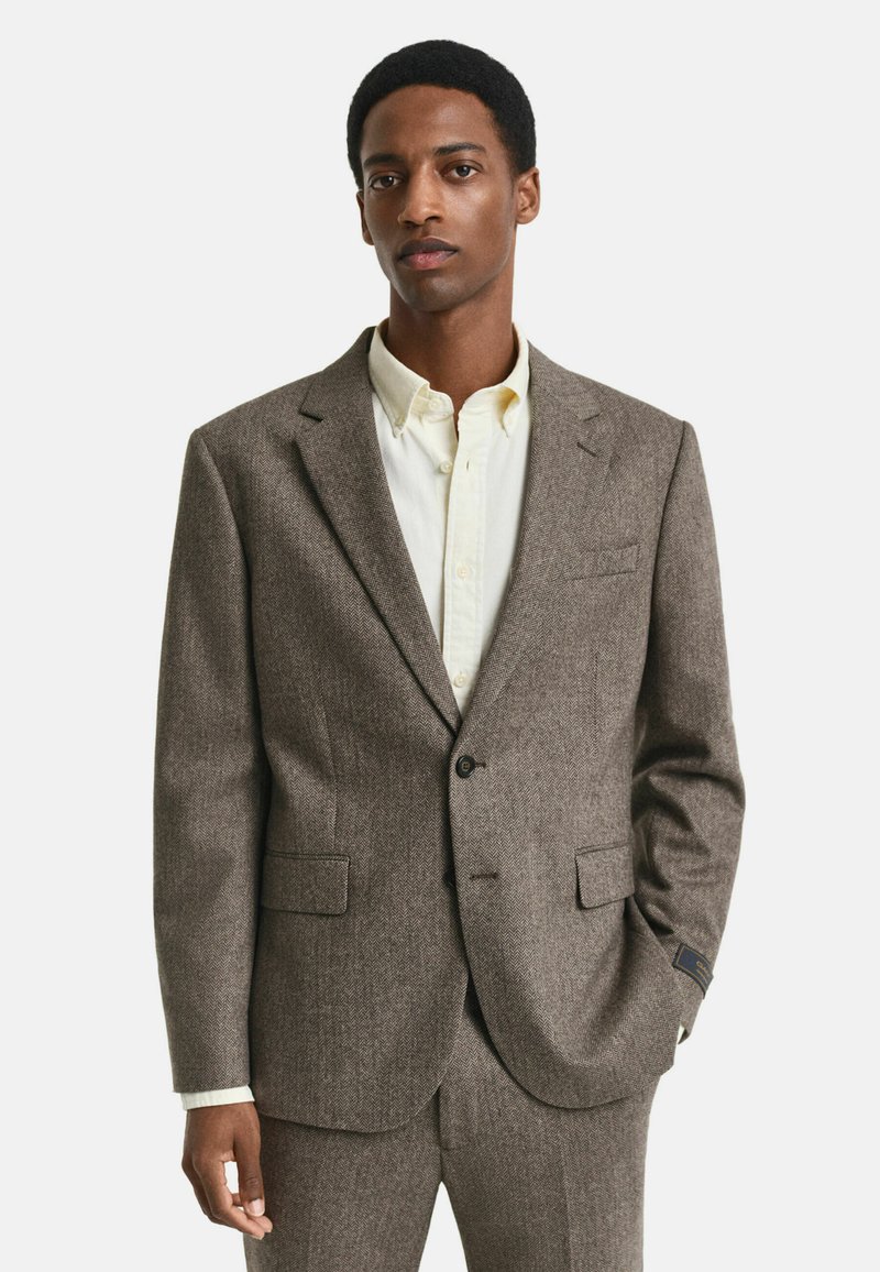 Giacca da abito a zigzag marrone con rever a punte, due tasche frontali e chiusura a un bottone. Indossata sopra una camicia di colore chiaro.