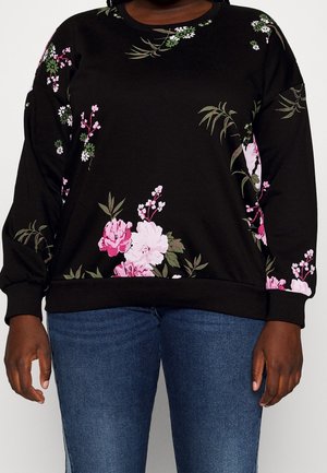 Sort langærmet top med pink og hvide blomsterprint, båret med blå jeans, vist fra nakke til hofter.