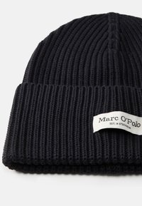 Gorro de punto negro con textura acanalada; cuenta con una etiqueta cosida que dice "Marc O'Polo" y "EST. EN ESTOCOLMO" en blanco.