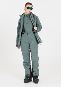 Tekniskt snöoutfit i dämpad grön, med en zip-up jacka, en åtsittande baslager och byxor med hängslen och svarta accenter.