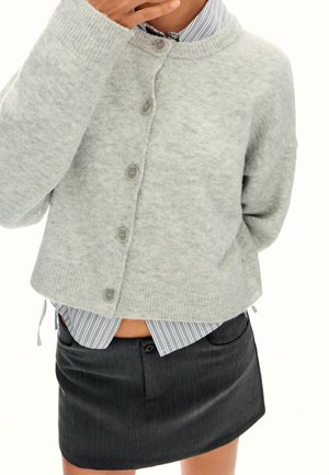 Personne portant un cardigan gris clair boutonné sur une chemise rayée rentrée dans une mini-jupe gris foncé, main près du visage.