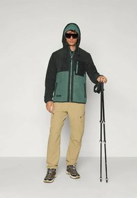 Fleecejacke in Dunkelgrün und Schwarz, mit Frontreißverschluss, verstellbarer Kapuze, Brusttasche; kombiniert mit beigen Cargo-Hosen und Trekkingstöcken.