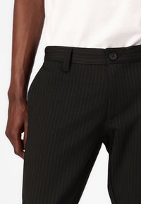 Pantalons à rayures noires ajustés, avec une fermeture à boutons, des passants de ceinture et une texture lisse. Les pantalons sont portés avec une chemise blanche.