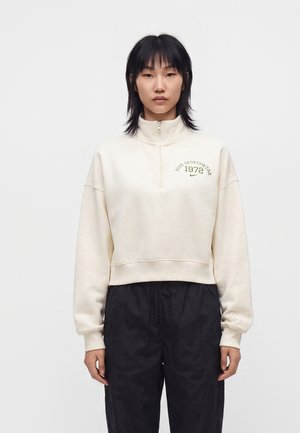 Ung person med lige sort hår bærer en cremefarvet sweatshirt med kvart lynlås med "Nike Sportswear 1972"-logo og sorte bukser, der står mod hvid baggrund.