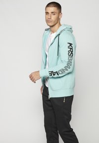 Sudadera con cremallera de color azul claro, con bolsillos laterales, que presenta una marca negra en las mangas y una capucha con cordón. Combinada con pantalones jogger negros.