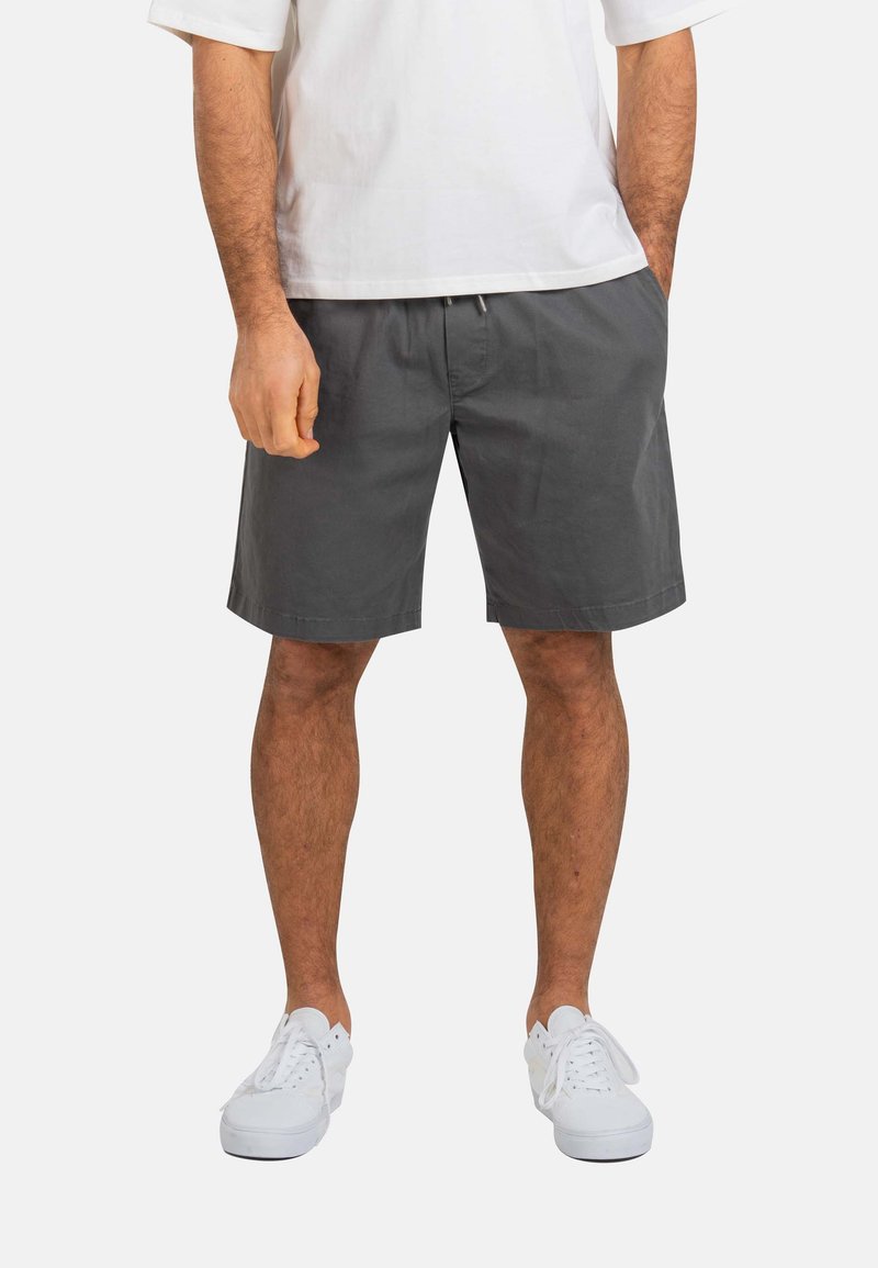 Hombre con pantalones cortos gris oscuro, zapatillas blancas y camisa blanca de manga corta, de pie con las manos en los bolsillos sobre un fondo liso.