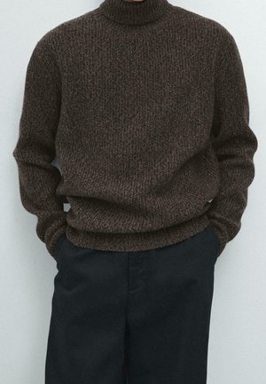 Person iført en mørkebrunt tekstureret rullekrave sweater og løstsiddende sorte bukser med hænderne i lommerne mod en ensfarvet baggrund.