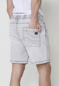 Pantalones cortos de mezclilla gris claro con cinturilla elástica, bolsillos laterales y un bolsillo trasero con parche que presenta una etiqueta de logo. Detalle de borde sin acabar.