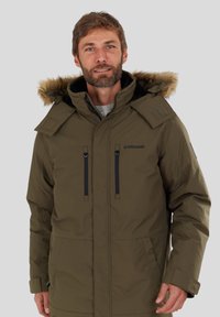 Uomo che indossa un parka invernale verde oliva con cappuccio foderato in pelliccia, tasche con zip nere e un maglione grigio sotto, in piedi contro uno sfondo neutro.