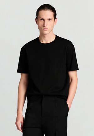 T-shirt nera a maniche corte realizzata in tessuto strutturato, con scollo rotondo e vestibilità rilassata, abbinata a pantaloni neri su misura.