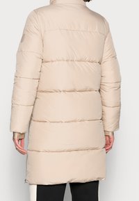 Abrigo acolchado beige con costuras horizontales, cuello alto y bolsillos con cremallera, que presenta una tela suave y brillante y una longitud hasta el muslo.
