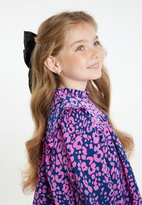 Blusa da bambino di colore blu navy con colletto arricciato, decorata con stampa leopardata rosa. Capelli lunghi acconciati con un fiocco nero.