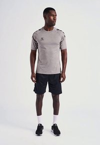 Maglietta sportiva grigia a maniche corte con accenti neri, abbinata a pantaloncini neri e scarpe nere. Il modello posa con sicurezza su uno sfondo neutro.